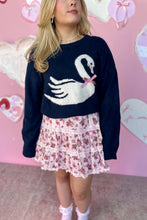 Load image into Gallery viewer, Person wearing a navy sweater with a white swan design against a pink background with heart shapes.