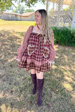 Load image into Gallery viewer, A girl wearing a brown plaid babydoll dress with heart pockets and brown boots standing in front of a white house with a brick fireplace.