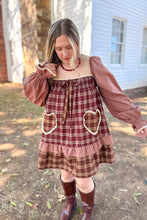 Load image into Gallery viewer, A girl wearing a brown plaid babydoll dress with heart pockets and brown boots standing in front of a white house with a brick fireplace.