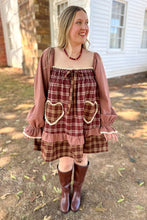 Load image into Gallery viewer, A girl wearing a brown plaid babydoll dress with heart pockets and brown boots standing in front of a white house with a brick fireplace.
