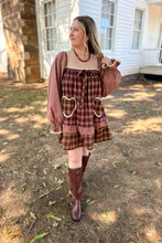 Load image into Gallery viewer, A girl wearing a brown plaid babydoll dress with heart pockets and brown boots standing in front of a white house with a brick fireplace.