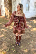 Load image into Gallery viewer, A girl wearing a brown plaid babydoll dress with heart pockets and brown boots standing in front of a white house with a brick fireplace.