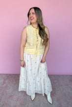 Load image into Gallery viewer, Woman wearing a yellow sleeveless top and white skirt with colorful embroidered floral pattern against a pink background