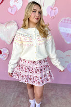 Load image into Gallery viewer, Woman wearing a cream cardigan and floral skirt against a pink background with heart shapes.