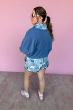Load image into Gallery viewer, Person wearing a blue jacket and floral shorts against a pink wall.