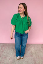 Load image into Gallery viewer, Woman wearing a green blouse and blue jeans against a pink background