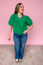 Load image into Gallery viewer, Woman wearing a green blouse and blue jeans against a pink wall.