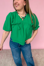 Load image into Gallery viewer, Woman wearing a green blouse with puff sleeves and blue jeans against a pink background