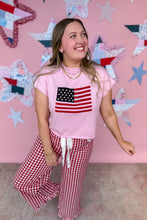 Load image into Gallery viewer, Woman wearing a pink t-shirt with an American flag design and red checkered pants against a pink wall with star decorations.