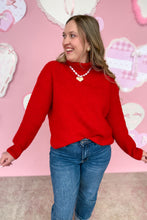 Load image into Gallery viewer, Woman wearing a red sweater and blue jeans against a pink background with heart decorations.