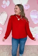 Load image into Gallery viewer, Woman wearing a red sweater and blue jeans standing in front of a pink wall with heart decorations.
