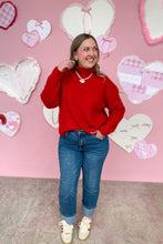 Load image into Gallery viewer, Person wearing a red sweater and blue jeans standing in front of a pink wall with heart decorations.