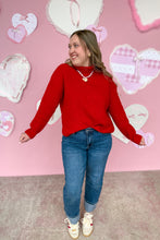 Load image into Gallery viewer, Woman wearing a red sweater and blue jeans standing in front of a pink wall with heart decorations.
