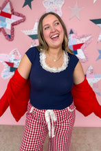 Load image into Gallery viewer, Woman wearing a navy top with lace trim and red checkered pants, standing in front of a pink wall with star decorations.