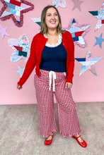 Load image into Gallery viewer, Woman wearing a red cardigan, navy top, and red and white checkered pants against a pink wall with star decorations.