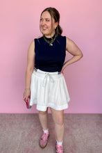 Load image into Gallery viewer, Model is wearing a navy blue mockneck and white skort against a pink background.