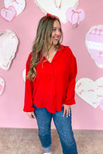 Load image into Gallery viewer, Woman wearing a red blouse and blue jeans standing in front of a pink wall with heart decorations.