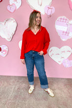 Load image into Gallery viewer, Woman in a red shirt and blue jeans standing in front of a pink wall with heart decorations.