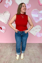 Load image into Gallery viewer, Woman in a red sleeveless top and blue jeans standing in front of a pink wall with heart decorations.