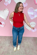 Load image into Gallery viewer, Woman wearing a red sleeveless top and blue jeans standing in front of a pink wall with heart decorations.