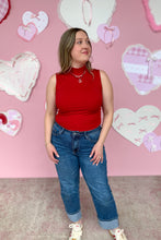 Load image into Gallery viewer, Woman wearing a red sleeveless top and blue jeans standing in front of a pink wall with heart decorations.