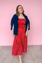 Load image into Gallery viewer, A woman modeling a red and white vertically striped midi dress with a smocked bodice, styled with a navy blue cardigan and beige strappy heels, set against a solid pink studio background.