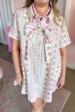 Load image into Gallery viewer, A woman wearing a short-sleeve pink and white floral print dress with a large bow at the collar, standing in a pink-themed room.