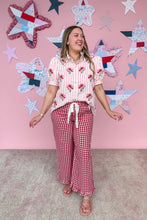 Load image into Gallery viewer, Woman wearing a floral top and red checkered pants against a pink wall with star decorations.