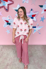 Load image into Gallery viewer, Woman wearing a floral top and red checkered pants against a pink wall with star decorations.