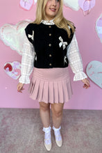 Load image into Gallery viewer, Person wearing a black cardigan vest with white bows and a pink pleated skirt against a pink background.
