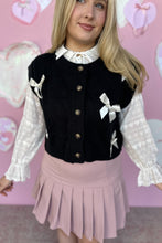 Load image into Gallery viewer, Person wearing a black cardigan vest with white bows, white blouse, and pink pleated skirt against a pink heart-patterned background.

