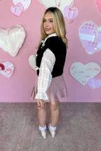 Load image into Gallery viewer, Woman in a black cardigan vest and white top with pink skirt with a pink heart-themed background
