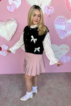 Load image into Gallery viewer, Woman wearing a black sweater vest with white bows, white blouse, pink pleated skirt, and white shoes in front of a pink heart-patterned wall.
