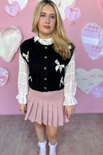Load image into Gallery viewer, Person wearing a black cardigan vest with white bow details, white blouse, and pink pleated skirt against a pink heart-patterned background.

