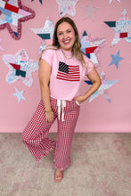 Load image into Gallery viewer, Woman wearing a pink t-shirt with an American flag design and red checkered pants against a pink wall with star decorations.
