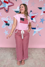 Load image into Gallery viewer, Woman wearing a pink t-shirt with an American flag design and red checkered pants against a pink wall with star decorations.
