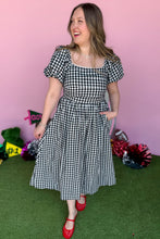 Load image into Gallery viewer, Girl wearing a black and white gingham midi dress with red jelly flats in front of a pink game day backdrop.
