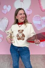 Load image into Gallery viewer, Woman wearing a sweater with a teddy bear design, holding a heart-shaped box and teddy bear, against a pink wall with heart decorations.
