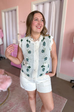 Load image into Gallery viewer, A woman modeling a sleeveless white scallop-trimmed vest with green floral patterns over a white top, paired with white shorts and beige flat shoes, in a pink boutique setting.
