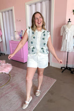 Load image into Gallery viewer, A woman modeling a sleeveless white scallop-trimmed vest with green floral patterns over a white top, paired with white shorts and beige flat shoes, in a pink boutique setting.
