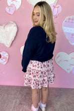 Load image into Gallery viewer, Woman wearing a navy sweater and floral skirt against a pink wall with heart decorations.
