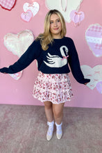 Load image into Gallery viewer, Woman wearing a navy sweater with a white swan design and a floral skirt against a pink background with heart shapes.
