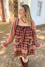 Load image into Gallery viewer, A girl wearing a brown plaid babydoll dress with heart pockets and brown boots standing in front of a white house with a brick fireplace. 
