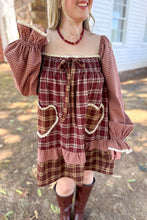 Load image into Gallery viewer, A girl wearing a brown plaid babydoll dress with heart pockets and brown boots standing in front of a white house with a brick fireplace. 
