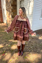 Load image into Gallery viewer, A girl wearing a brown plaid babydoll dress with heart pockets and brown boots standing in front of a white house with a brick fireplace. 

