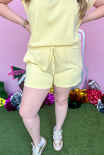 Load image into Gallery viewer, Girl wearing a yellow set with matching shorts and short sleeve top with a pink backdrop. 
