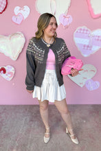 Load image into Gallery viewer, Woman wearing a dark gray patterned cardigan over a pink top and white skirt, standing against a pink wall with heart decorations.

