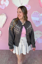 Load image into Gallery viewer, Woman wearing a dark gray cardigan with white patterns, pink top, and white skirt against a pink wall with heart decorations.

