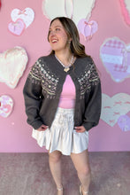 Load image into Gallery viewer, Woman wearing a dark gray cardigan with heart and bow details, pink top, and white skirt against a pink background with heart shapes.
