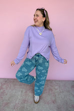 Load image into Gallery viewer, Woman wearing a purple long-sleeve top and blue floral pants against a pink wall.
